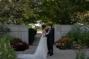 A wedding couple poses in front of a white wall surrounded by lush garden greenery.