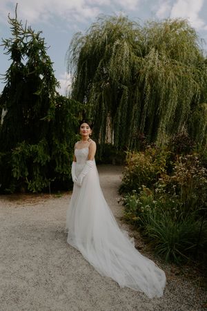 Flowing white wedding dress trails behind on garden path near weeping willow trees.