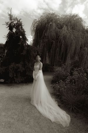 Black and white photo of wedding dress flowing on garden path near willow trees.