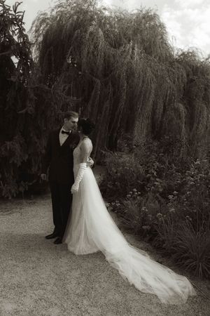 An elegant black and white wedding photo featuring a flowing dress train beneath weeping willow branches.