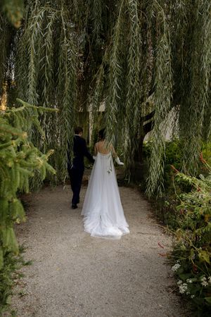 A wedding couple walks through a garden path lined with drooping willow branches.