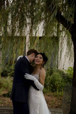 A couple embraces and laughs together during an outdoor wedding photo session beneath drooping willow branches.