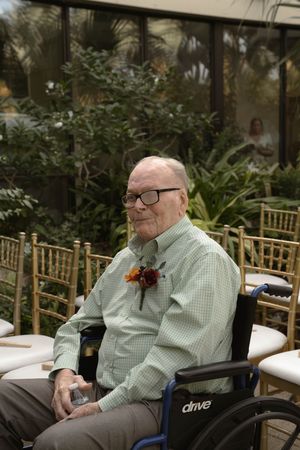 An elderly person in a wheelchair wearing a light green shirt sits in a garden setting with bamboo chairs.