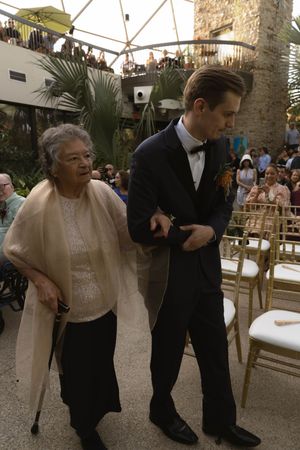 Formal event attendees assist elderly guests with walking at an outdoor venue.