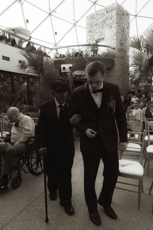 Formal event attendees assist elderly guests walking through an outdoor venue.
