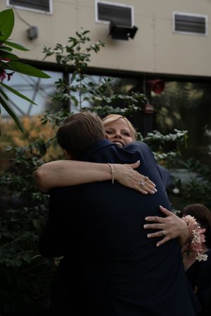An emotional embrace between two people at a formal event with greenery in the background.