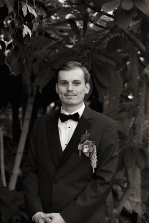 A formal portrait of someone in a black tuxedo with bow tie and boutonniere.