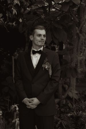 A groom in a classic black tuxedo standing in a garden setting.