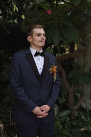 A groom in formal attire with boutonniere poses in front of lush garden foliage.