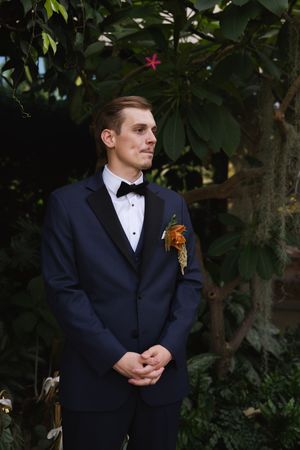 A contemplative groom in formal attire standing among garden greenery.