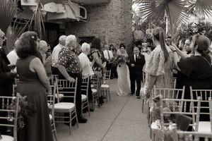 Wedding guests gather in an elegant courtyard venue decorated with palm trees and chiavari chairs for a ceremony.