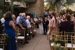 Outdoor wedding ceremony setup with gold chairs and tropical foliage at a brick-walled venue.