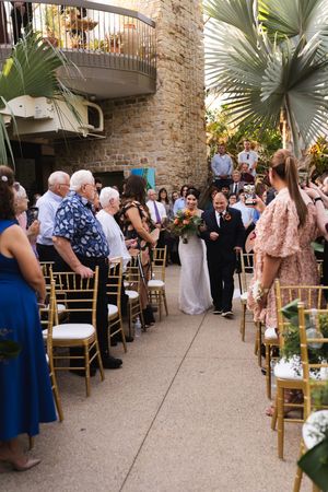 Wedding processional in a tropical courtyard with guests standing along the aisle.