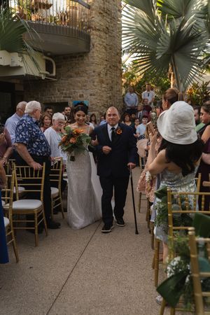 Wedding ceremony guests standing in a courtyard with palm trees and brick walls.