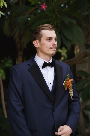 Formal portrait of a groom in a navy tuxedo with an orange boutonniere against a dark background.