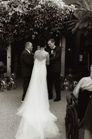 A flowing white wedding dress trails behind as people walk through a garden setting.