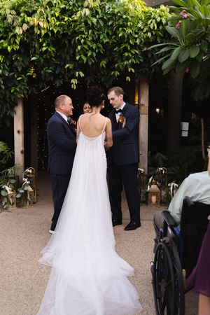 A wedding ceremony takes place in a lush garden setting with flowing white dress and formal attire.