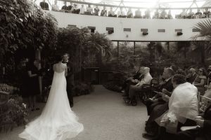An elegant wedding ceremony in a conservatory with string lights and tropical foliage overhead.