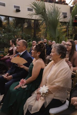 Wedding guests in formal attire smile and react during an indoor garden ceremony.