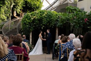 Wedding ceremony in progress at an outdoor garden venue with greenery and white decor.