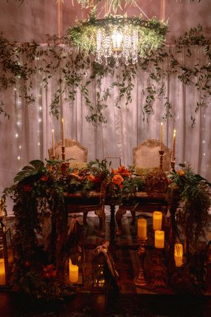 An enchanting sweetheart table with hanging greenery, candles, and elegant chairs under a crystal chandelier.