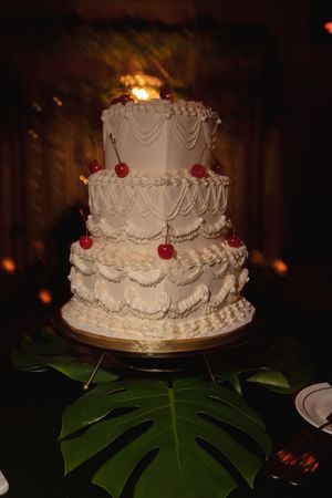 Classic white tiered wedding cake with intricate piping and red decorative elements on a green leaf base.