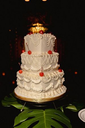 A classic white wedding cake with delicate piping and cherry decorations on a dark background.