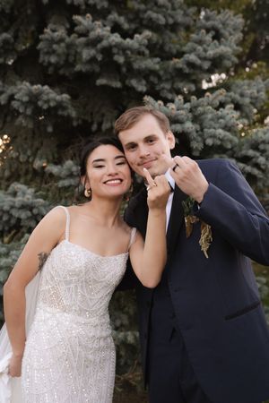A couple poses together outdoors in wedding attire with blue spruce trees in the background.