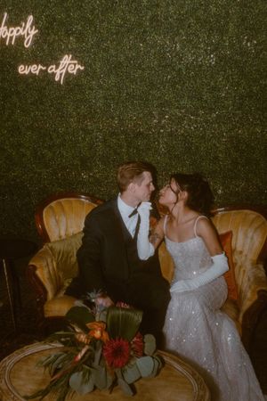 Newlyweds share romantic moments on vintage chairs against a green backdrop with illuminated text.