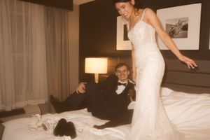 A couple in formal attire poses playfully on a hotel bed with black and white artwork on the wall.