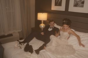 A romantic wedding photo featuring a couple lounging on white bedding with wall art and a table lamp.