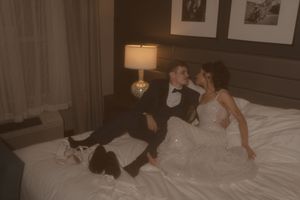 A warm-toned wedding portrait showing a couple in formal attire on white hotel bedding.