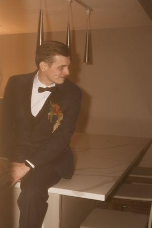 A groom in formal attire leans against a counter in warm ambient lighting.