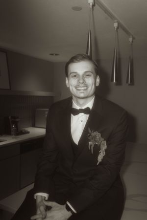 Person in formal black tuxedo and bowtie smiles while seated in dimly lit room.