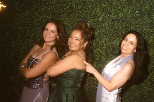 Three women in formal dresses pose playfully against a green hedge wall at an evening event.