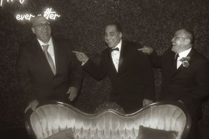 Black and white photo of three men in formal wear gesturing behind an antique sofa.