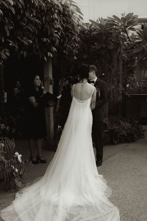 Flowing white wedding dress with delicate details photographed in an outdoor garden setting.