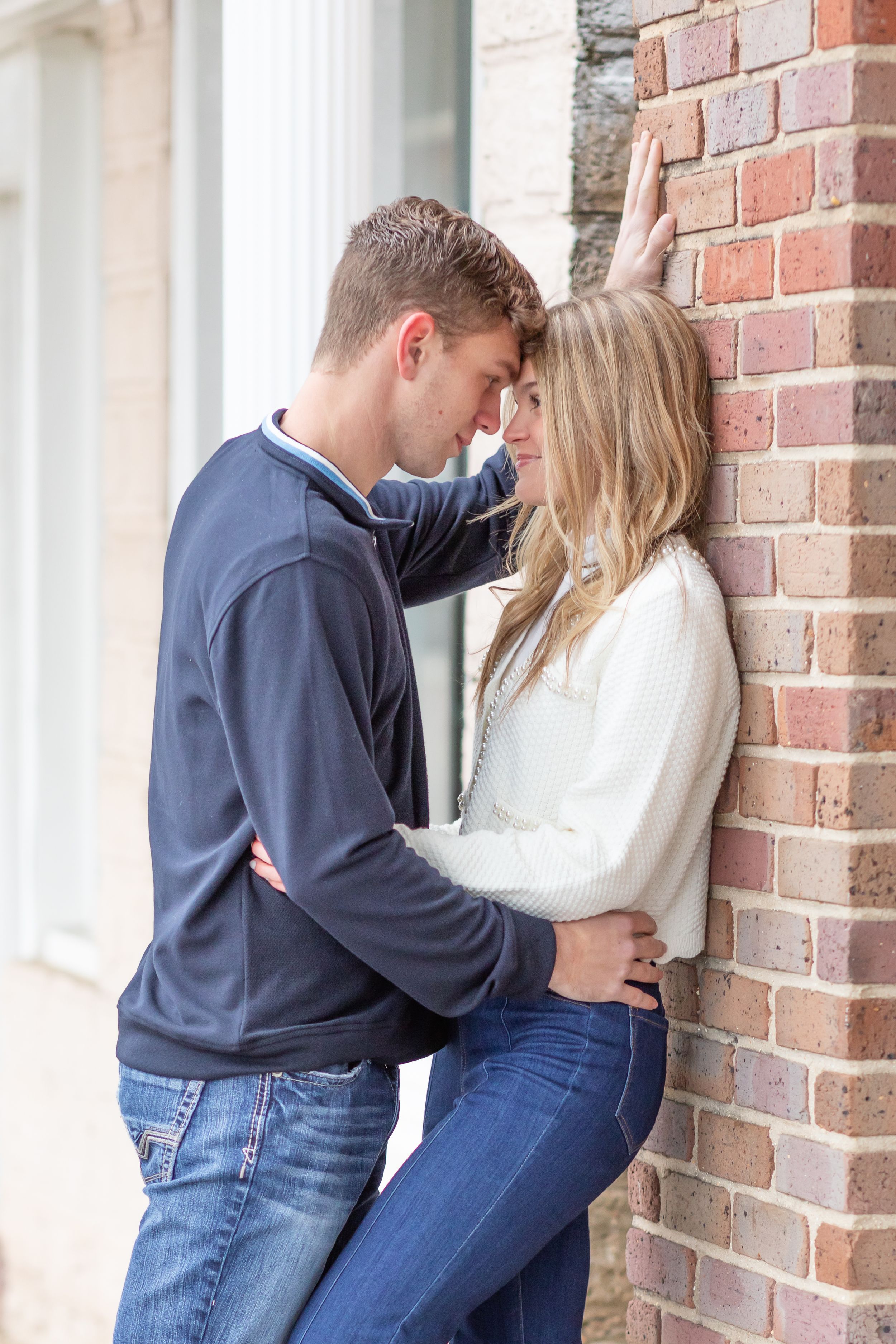 Romantic couple poses against brick wall, wearing coordinated casual outfits.
