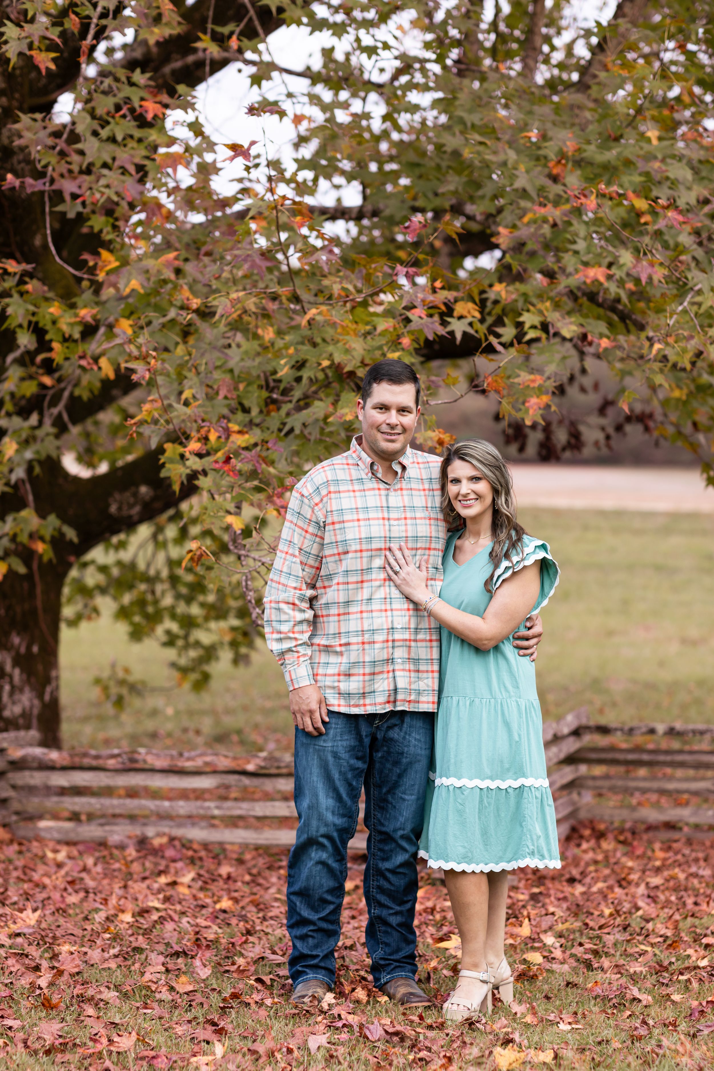 Couple poses together in autumn setting with fallen leaves and trees.