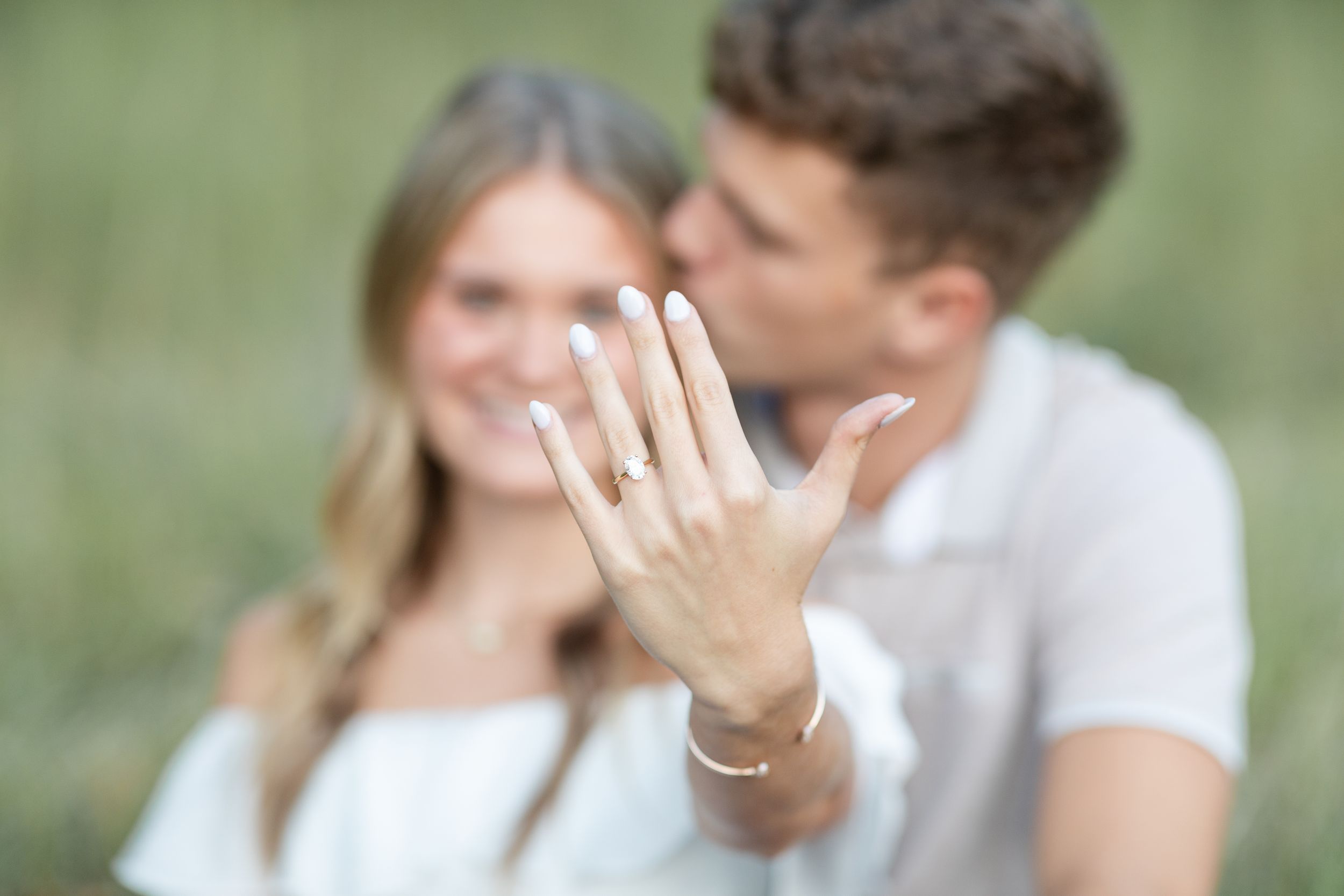 Hand displaying engagement ring in soft focus with couple in background.