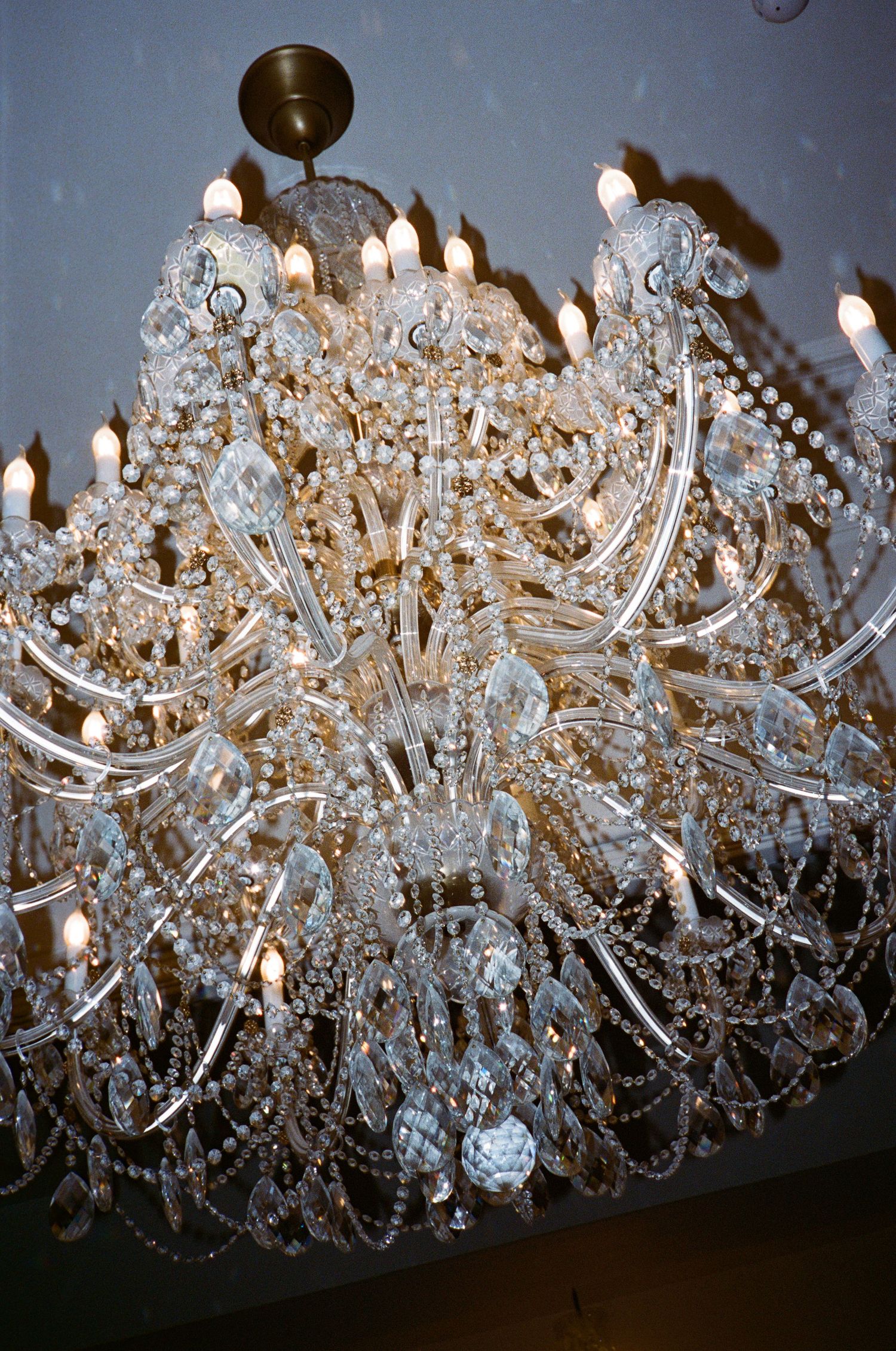Elegant crystal chandelier with multiple tiers of sparkling glass drops and lit candle-style bulbs illuminates a dark space.