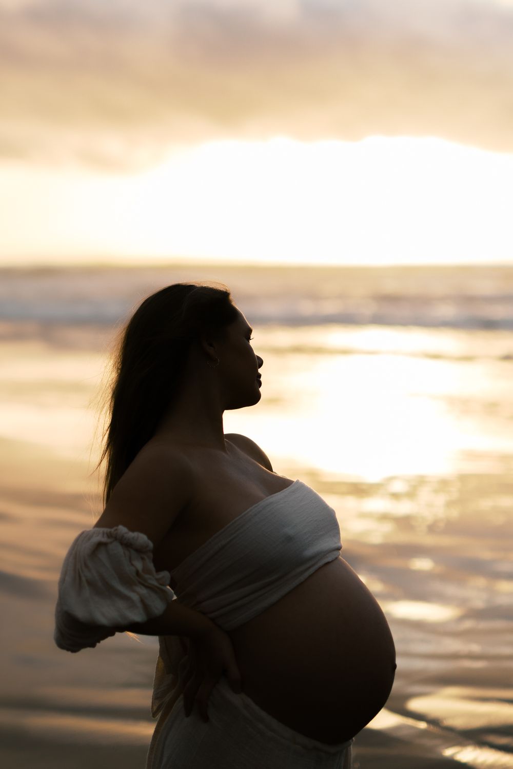 Silhouette of an expectant mother in white dress against a golden sunset at the beach.