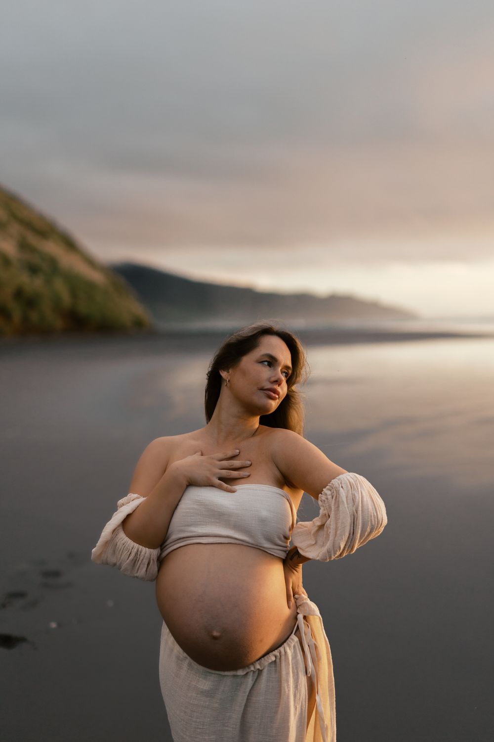 Dramatic coastal maternity portrait with mountains and sunset sky in background.