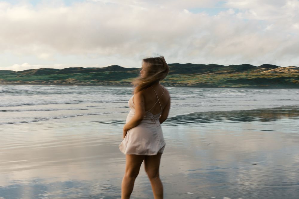 A person in a white dress stands on a sandy beach with windswept hair as waves roll in at sunset.