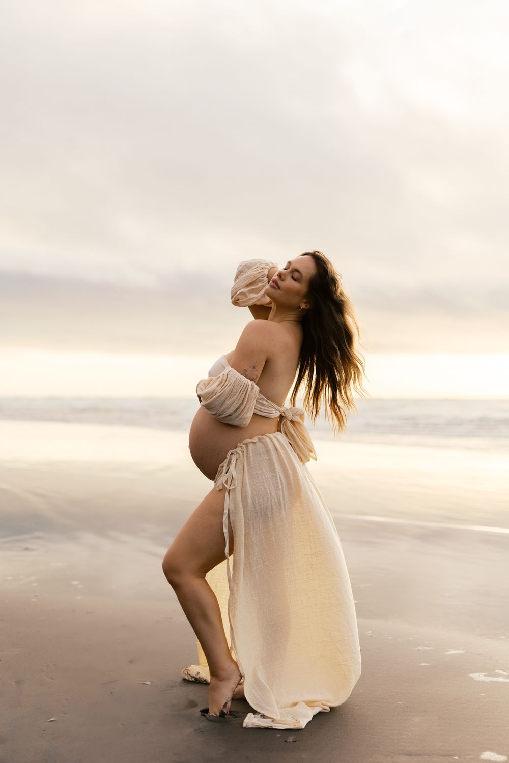 Artistic maternity photo session at sunset on a beach with flowing white dress and natural lighting.