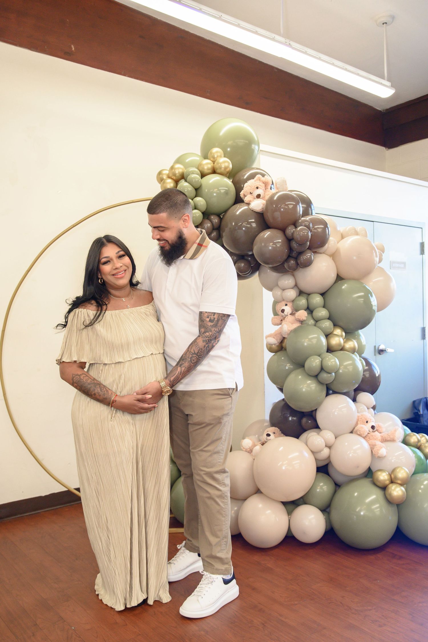 A romantic maternity photoshoot with a couple posing against a neutral backdrop with sage green and white balloon arch decor.