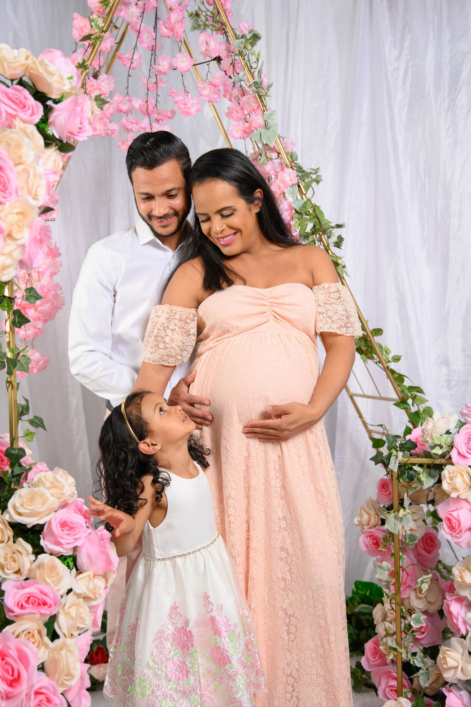 Romantic maternity photo session with couple in white and peach outfits surrounded by pink floral decorations.