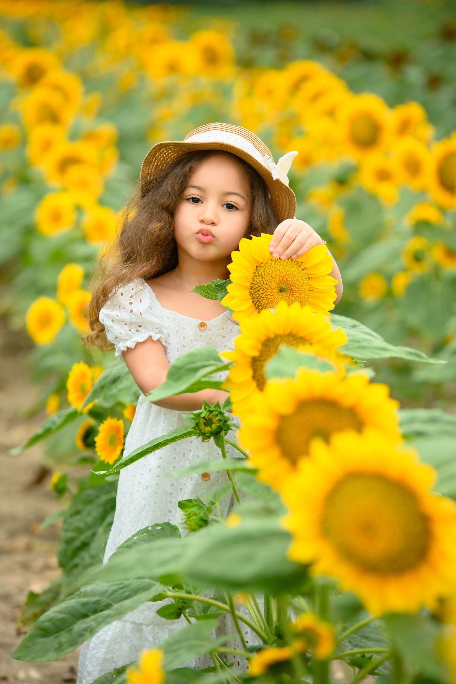 A child in a white dress poses playfully among bright yellow sunflowers in a garden setting.