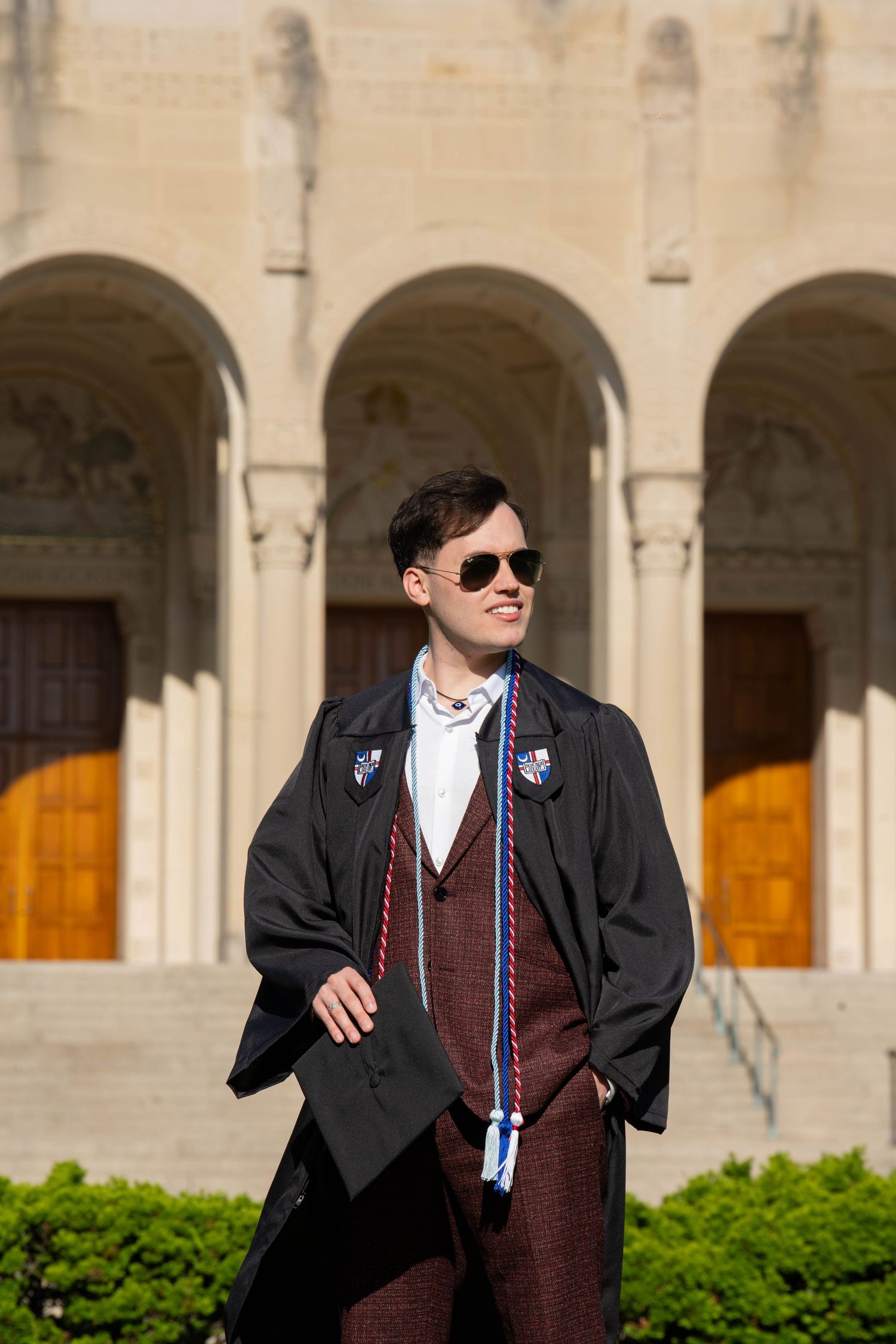 Graduate in black cap and gown poses with playful gestures in front of arched university building.
