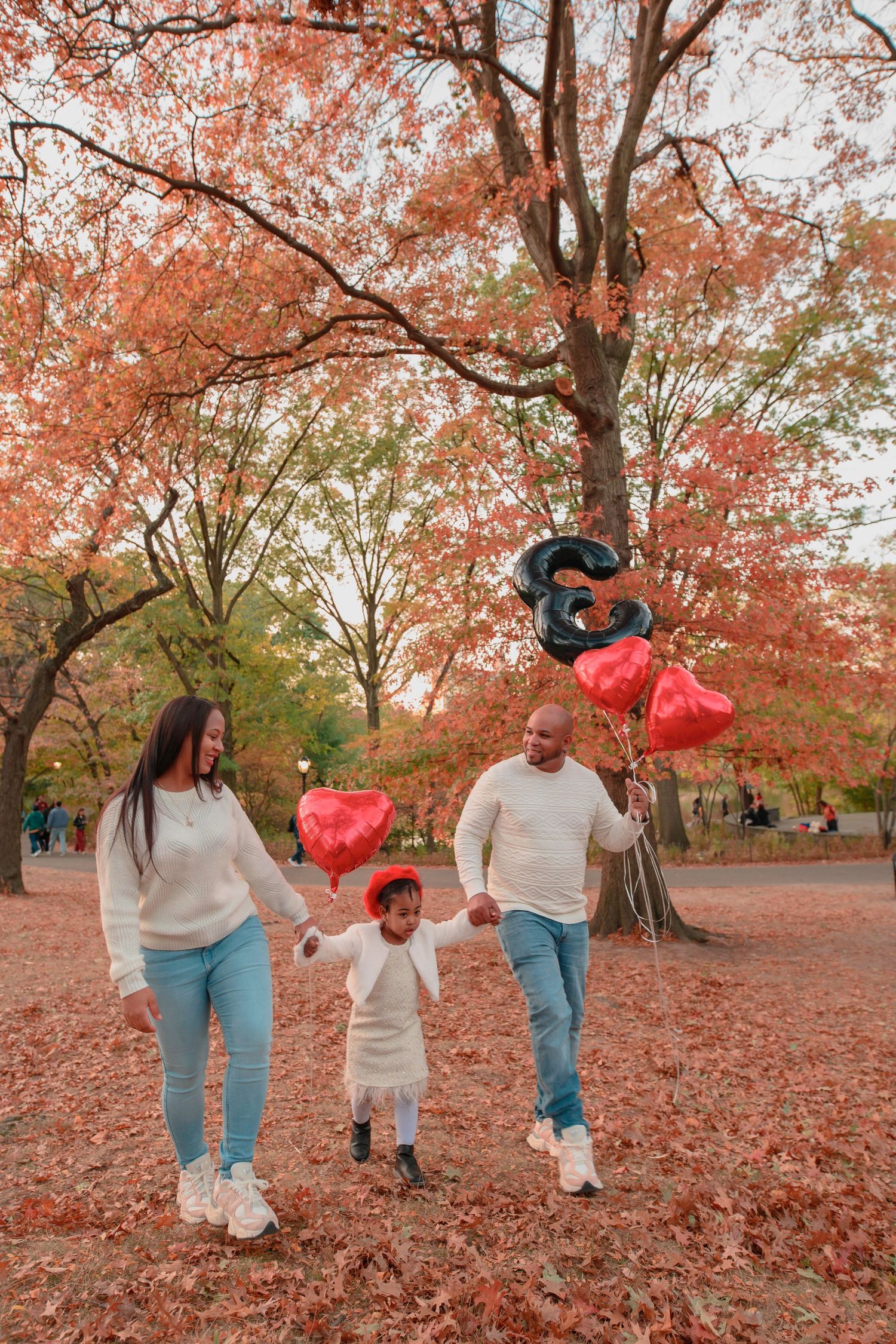 Family walking together through autumn park with red balloons and fallen maple leaves on ground in warm sunlight.